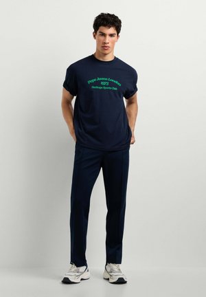 Mann trägt ein dunkelblaues Pepe Jeans London 1973 Heritage Sports Club T-Shirt, dunkelblaue Hose und weiße Turnschuhe, steht vor einer hellen Wand.