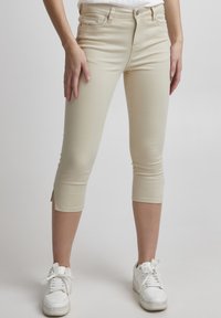 Beige capri bikses, izgatavotas no elastīgas auduma, ar pieguļošu dizainu, potītes garumu un nelieliem sānu slitēm. Valkātas ar baltām kedām.