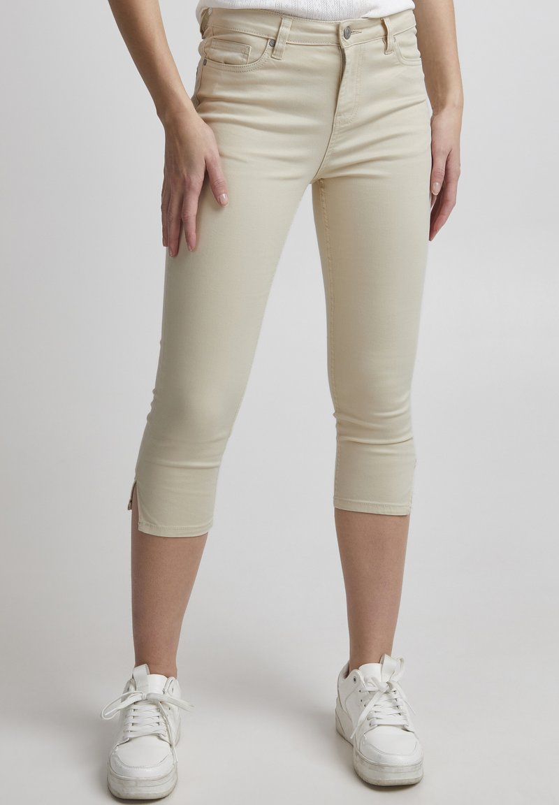 Beige capri bikses, izgatavotas no elastīgas auduma, ar pieguļošu dizainu, potītes garumu un nelieliem sānu slitēm. Valkātas ar baltām kedām.