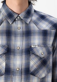 Camicia di flanella a quadri blu e crema con colletto a punta, bottoni a scatto, due tasche sul petto e un'etichetta con il logo Wrangler sulla tasca sinistra.