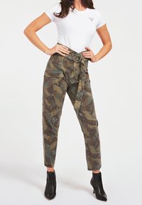 Hoge camouflagebroeken in tinten groen en bruin, met een riem, zakken en een losse pasvorm. Gepaard met een wit T-shirt.