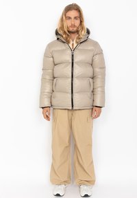 Pufferjack in lichtbeige, met een capuchon, ritssluiting aan de voorkant en elastische manchetten, gecombineerd met losse beige cargobroeken en sneakers.