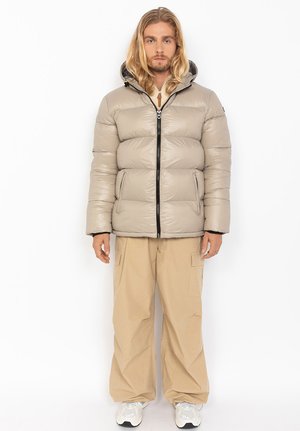 Pufferjack in lichtbeige, met een capuchon, ritssluiting aan de voorkant en elastische manchetten, gecombineerd met losse beige cargobroeken en sneakers.