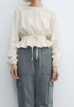 Kobieta ubrana w białą bluzkę z długim rękawem, zdobioną koronką i ściągaczem w talii, zestawioną z szarymi, wysokim stanem spodniami cargo.