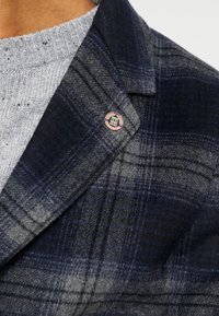 Cappotto di lana a quadri navy e grigio con colletto a doppio, indossato sopra un maglione lavorato a maglia grigio chiaro, con un dettaglio a spilla smaltata circolare.