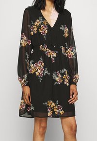 Černé květinové šaty s V výstřihem, dlouhými průhlednými rukávy, nařaseným pasem a pestrými květinovými vzory v odstínech žluté, růžové a zelené.