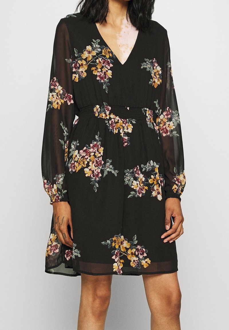 Černé květinové šaty s V výstřihem, dlouhými průhlednými rukávy, nařaseným pasem a pestrými květinovými vzory v odstínech žluté, růžové a zelené.