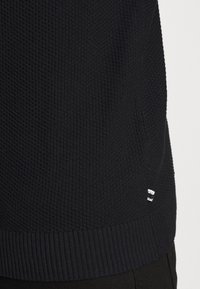 Mörkstickad tröja med ett texturerat mönster, ribbad nederkant och en liten logotyp i vitt. Designen betonar värme och komfort.