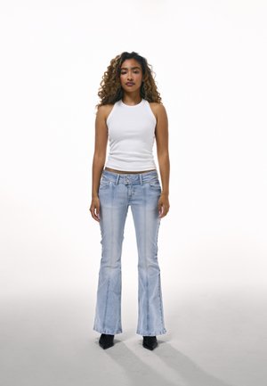 Kobieta o kręconych włosach stoi zwrócona twarzą do przodu, ubrana w biały, bezrękawnik, jasnoniebieskie rozszerzane jeansy i czarne, spiczaste buty.