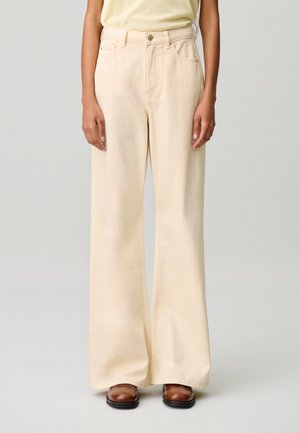 Femme portant un pantalon large beige, un haut jaune clair et des chaussures marron, debout devant un fond clair uni.
