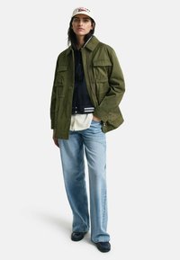 Olivgrüne Jacke mit mehreren Fronttaschen, über einem dunkelblauen Hemd und einer weißen Bluse getragen, kombiniert mit hellblauen Jeans und einer Mütze.