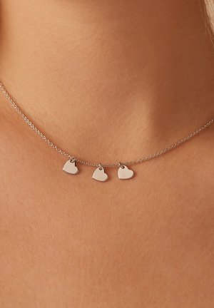 Zilveren halsketting met drie kleine hartbedels, met een delicate ketting en een glad oppervlak, rustend op een huidkleurige achtergrond.