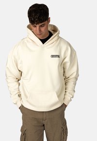 Felpa color crema realizzata in tessuto morbido, dotata di una tasca frontale. Presenta un ampio cappuccio e la parola "CRYSTAL" in testo nero sul petto.
