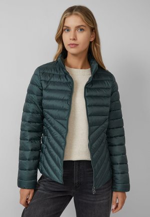Pufferjack in donker tealkleur, met een ritssluiting en een chevron-gequilt patroon. Gecombineerd met een crèmekleurige trui en zwarte jeans.