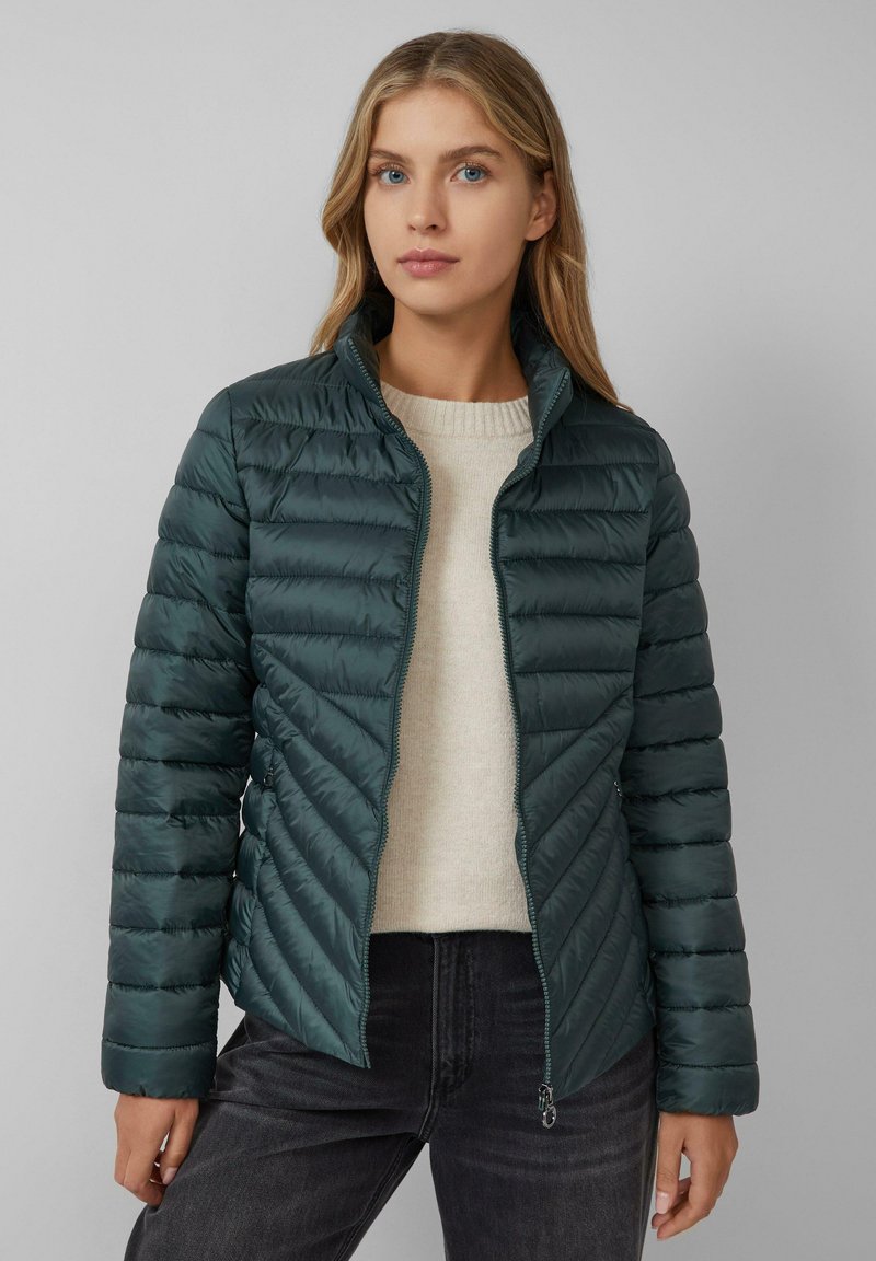 Doudoune en teal foncé, avec fermeture éclair et motif matelassé en chevrons. Associée à un pull crème et un jean noir.