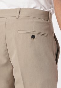 Beige tailored trousers with a smooth texture, featuring a side pocket with a black button and minimalist design.