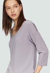 Helllila V-Ausschnitt-Top aus strukturiertem Stoff, mit Dreiviertelärmeln und einem kleinen Markenetikett am Saum.