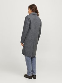 Cappotto di lana grigio con colletto alto, tessuto leggermente strutturato, di lunghezza media e taglio dritto. Indossato sopra jeans blu larghi e scarpe nere.