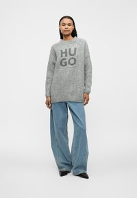 Gray knitted sweater with oversized fit, featuring "HUGO" logo in darker gray. Paired with light blue wide-leg jeans and black shoes.