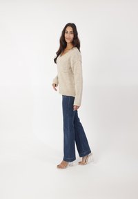 Beige gestrickter Pullover mit V-Ausschnitt und frontaler Tasche, kombiniert mit dunkelblauen Schlagjeans und transparenten Absatzsandalen. Einheitlicher Hintergrund.