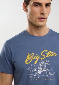 Zila kokvilnas t-kreklu ar dzeltenu "Big Star" grafikā, kurā redzama zirdziņa un jātnieka ilustrācija, un apakšā ir teksts "KOPŠ 1970".