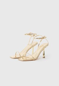 Sandales à talons hauts dorées avec un design à bout carré, des fines lanières, une fermeture à boucle à la cheville et une finition métallique brillante.