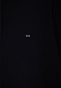 Pull noir en maille avec un petit logo rose sur le côté gauche. La texture est lisse et le tissu semble dense.