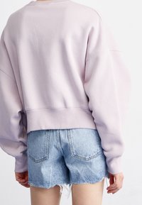 Osoba ubrana w jasnofioletową, oversize'ową bluzę oraz jasnoniebieskie, wysokowciskowe szorty z denimu z postrzępionymi krawędziami, widziana od tyłu.
