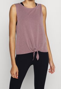 Haut sans manches mauve texturé avec un nouage à l'avant porté sur un brassière de sport noire, associé à un legging noir ajusté sur une personne debout.
