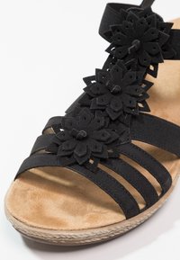 Sandale noire avec trois lanières à l'avant, bande élastique à la cheville, semelle rembourrée beige et appliques florales noires décoratives au centre.