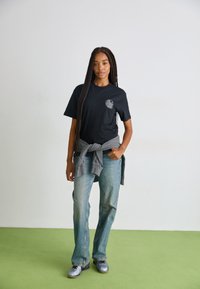 Svart bomullströja med grafisk design på vänster bröst, parat med ljusblå utsvängda jeans och grå tröja knuten i midjan.