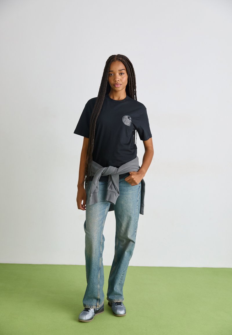 Zwarte katoenen t-shirt met een grafisch ontwerp op de linkerborst, gecombineerd met lichtblauwe uitlopende jeans en een grijze trui die om de taille is geknoopt.
