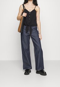 Černý top bez rukávů s knoflíky a detailem uzlu vpředu, zkombinovaný s širokými tmavými džíny a černými platformovými botami. Hnědá taška přes rameno.