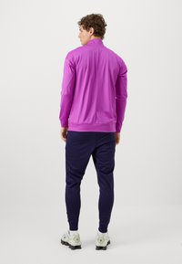 Paarse sportjack met een staande kraag, gedragen met marineblauwe joggingsbroeken en witte sneakers. Gladde textuur, aansluitend ontwerp en elastische manchetten.