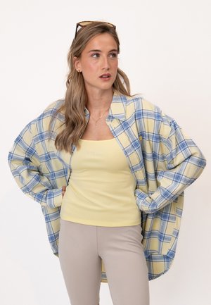 Jeune femme aux longs cheveux blonds porte des lunettes sur la tête, un débardeur jaune, une chemise à carreaux jaune et bleue oversize et un pantalon beige, mains sur les hanches.