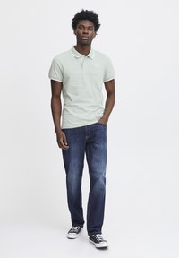 Polo shirt verde chiaro con colletto e logo, abbinato a jeans blu scuro e sneakers in tela nere. Il modello è in posizione frontale.