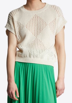 Femme portant un pull en maille crème à manches courtes avec motif losange et une jupe plissée verte, montrée des épaules aux hanches.