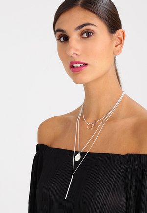 Zilveren gelaagde kettingen met een ronde hanger, een ketting met een kleine schijf en een verticale staafaccent. Draagt over een off-shoulder zwarte top.