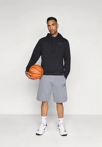 Črna kapucar s kapuco z logotipom, sive športne kratke hlače, drži košarkarsko žogo, dopolnjeno s belimi supergami in nogavicami. Enobarvno ozadje.