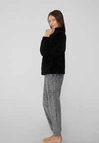 Zwarte, fluffy jas met een hoge kraag, gecombineerd met grijze pyjamabroek met een subtiel hartpatroon. Model staat blootsvoets in zijaanzicht.