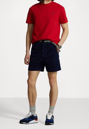 Man draagt een rood shirt met korte mouwen, marineblauwe korte broek met een gestreepte riem, grijze sokken en blauwe sportschoenen, staat met één hand in de zak.
