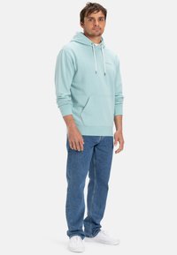 Mint green hooded sweatshirt with a front pocket, drawstring, and ribbed cuffs, paired with blue denim jeans and white sneakers.