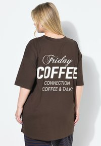 Brun oversized t-shirt lavet af bomuld med hvid tekst på bagsiden: "Fredag KAFFE FORBINDING 'KAFFE & SAMTALE'." Korte ærmer.