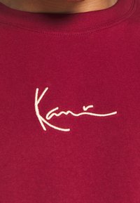 Burgundské bavlněné tričko s bílou vyšívanou signaturou umístěnou na prsou. Hladká textura a kulatý výstřih.