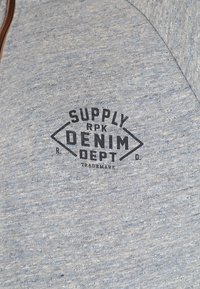 Gray-blue zip-up sweatshirt with a textured fabric. Features a printed logo "SUPPLY RPK DENIM DEPT" in black. Brown zipper accent.