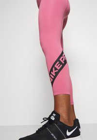 Leggings deportivos rosas con una franja diagonal negra que presenta el texto "NIKE PRO". Combinados con zapatillas negras. Tejido suave y elástico.