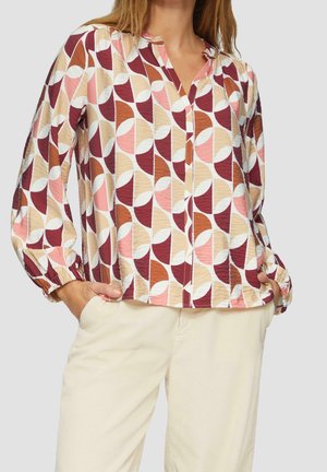 Frau trägt eine langärmelige Bluse mit einem rosa-, braun- und beigen geometrischen Muster, die in hellbeige Hosen gesteckt ist. Hände in den Taschen.