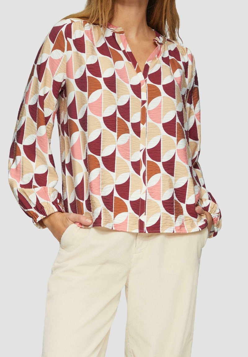 Femme portant un chemisier à manches longues avec un motif géométrique rose, marron et beige, rentré dans un pantalon beige clair. Mains dans les poches.