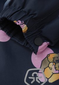 Marinblått tyg med fiskbensmönster, som har en tecknad lejon grafik och rosa cirkulära accenter. Elastisk mudd och svart tryckknapp.