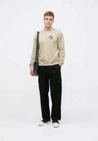 Beige pulóver fekete logóval, laza fekete cargo nadrággal, oldalsó zsebekkel párosítva. A fekete sportcipő teszi teljessé az öltözetet.